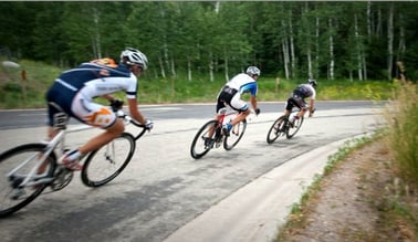 Bicyclers riding through a road with forest trees on the sides. 
